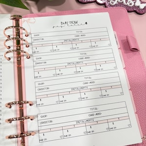 May include: A pink planner with rose gold hardware, open to a page with a "Buy Now, Pay Later" layout. The page has sections for shop, creditor, and installment amounts. A glittery sign that says "ezbudgets" is in the background.