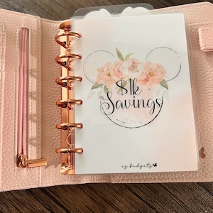May include: A pink and white floral design with a black outline of Mickey Mouse ears. The text "$1k Savings" is in the center of the design. The text "ez-budgets" is in the bottom right corner of the design.