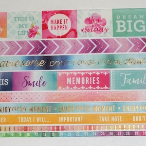 May include: A collection of colorful washi tapes with inspirational messages and gold accents. The tapes feature phrases like "Dream Big", "Make it Happen", "Love This", "Smile", and "Memories".