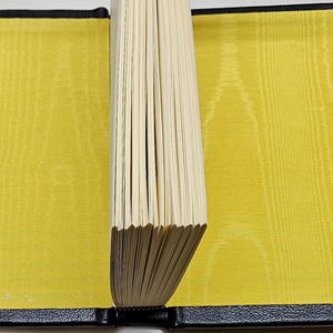 May include: An open book with a black leather cover and yellow silk-like pages. The pages are held together by a black spine. The inside covers are a bright yellow colour. The book is open, revealing the pages.
