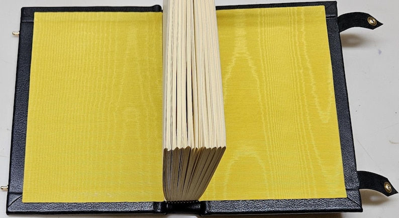 May include: An open book with a black leather cover and yellow silk-like pages. The pages are held together by a black spine. The inside covers are a bright yellow colour. The book is open, revealing the pages.