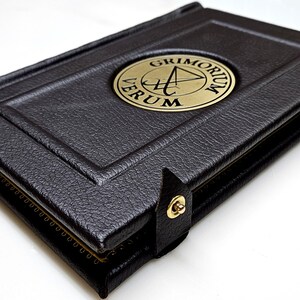 May include: A black leather-bound book with a gold embossed circle on the cover. The circle contains a symbol and the text "GRIMORIUM VERUM".