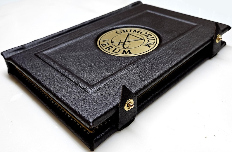 May include: A black leather-bound book with a gold embossed circle on the cover. The circle contains a symbol and the text "GRIMORIUM VERUM".