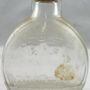 Puede incluir: Una botella de vidrio transparente y vintage con tap&oacute;n de corcho. La botella tiene el texto en relieve "HOLY WATER" y una forma redondeada. La botella est&aacute; ligeramente polvorienta, lo que sugiere su antig&uuml;edad. Una peque&ntilde;a mancha de color claro es visible en la superficie.