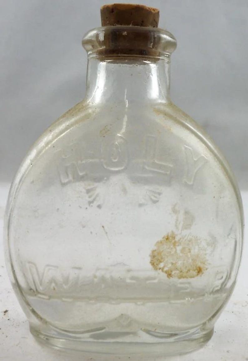 Puede incluir: Una botella de vidrio transparente y vintage con tap&oacute;n de corcho. La botella tiene el texto en relieve "HOLY WATER" y una forma redondeada. La botella est&aacute; ligeramente polvorienta, lo que sugiere su antig&uuml;edad. Una peque&ntilde;a mancha de color claro es visible en la superficie.