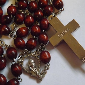 Puede incluir: Un rosario con cuentas de madera marr&oacute;n oscuro y una cruz de metal plateado. La cruz est&aacute; inscrita con "ROMA" e "ITALY". El rosario tambi&eacute;n presenta una pieza central plateada con una figura religiosa.