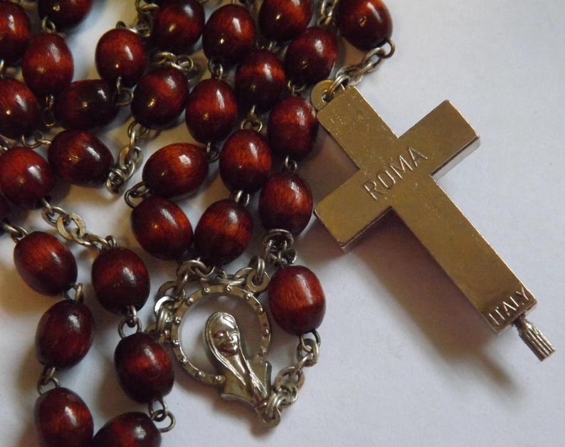 Puede incluir: Un rosario con cuentas de madera marr&oacute;n oscuro y una cruz de metal plateado. La cruz est&aacute; inscrita con "ROMA" e "ITALY". El rosario tambi&eacute;n presenta una pieza central plateada con una figura religiosa.