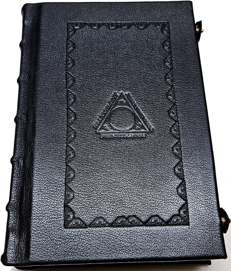 May include: A black leather-bound book with a textured cover. The cover features an embossed design with a triangle and circle symbol, and decorative border. The spine has raised bands. The book is closed with a metal clasp.
