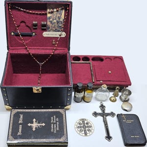 Puede incluir: Un estuche de viaje de cuero sint&eacute;tico negro con detalles dorados, abierto para revelar un interior burdeos y objetos religiosos. Incluye un rosario, un crucifijo, peque&ntilde;as botellas y un libro titulado "Roman Ritual".