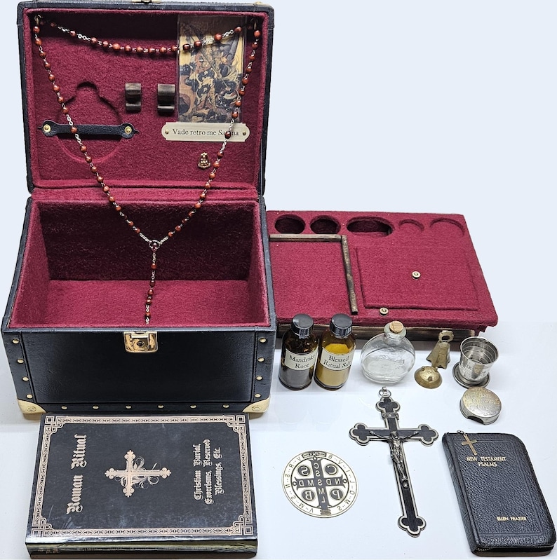 Puede incluir: Un estuche de viaje de cuero sint&eacute;tico negro con detalles dorados, abierto para revelar un interior burdeos y objetos religiosos. Incluye un rosario, un crucifijo, peque&ntilde;as botellas y un libro titulado "Roman Ritual".