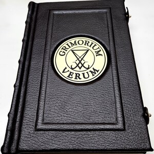 May include: A black leather-bound book with a gold embossed circle on the front cover. The circle contains the text "GRIMORIUM VERUM" and a symbol with a pentagram and two crossed swords.