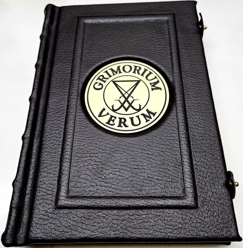 May include: A black leather-bound book with a gold embossed circle on the front cover. The circle contains the text "GRIMORIUM VERUM" and a symbol with a pentagram and two crossed swords.