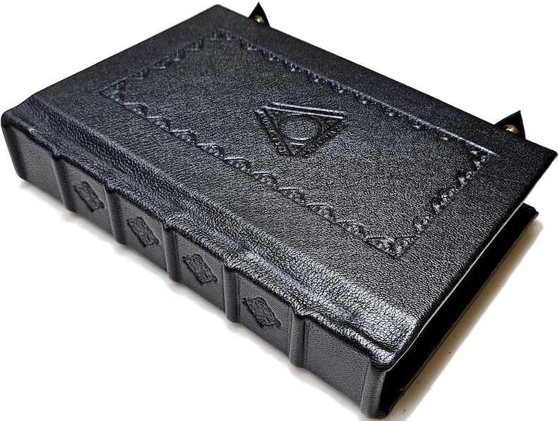 May include: A black leather-bound journal with embossed geometric designs and a triangular emblem on the cover. The spine features decorative panels. The journal has a textured surface and is closed with a small clasp.
