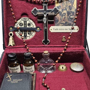 Puede incluir: Una caja negra con interior rojo, que contiene objetos religiosos. Incluye un crucifijo, rosario, peque&ntilde;as botellas etiquetadas como "Mandrake Root" y "Blessed Ritual Salt", un peque&ntilde;o libro y un medall&oacute;n de metal. La caja est&aacute; abierta.
