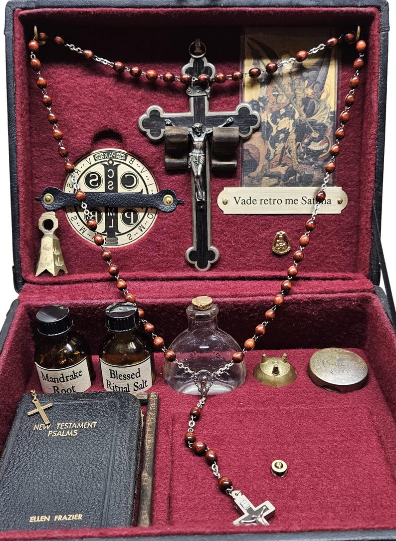 Puede incluir: Una caja negra con interior rojo, que contiene objetos religiosos. Incluye un crucifijo, rosario, peque&ntilde;as botellas etiquetadas como "Mandrake Root" y "Blessed Ritual Salt", un peque&ntilde;o libro y un medall&oacute;n de metal. La caja est&aacute; abierta.