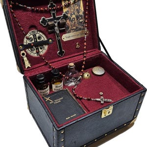 Puede incluir: Caja negra de estilo vintage con protectores de esquina dorados, abierta para revelar un interior de terciopelo rojo. Contiene un crucifijo, cuentas de rosario, un libro peque&ntilde;o, botellas y un medall&oacute;n. La caja se asegura con un pestillo dorado.