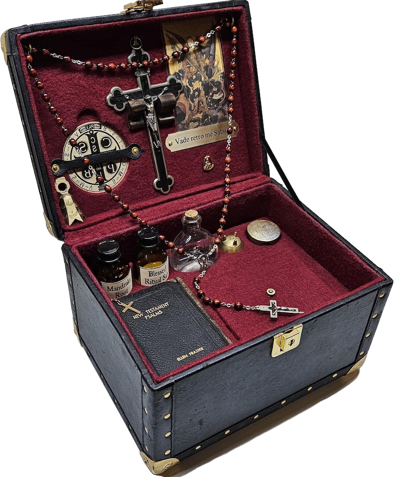 Puede incluir: Caja negra de estilo vintage con protectores de esquina dorados, abierta para revelar un interior de terciopelo rojo. Contiene un crucifijo, cuentas de rosario, un libro peque&ntilde;o, botellas y un medall&oacute;n. La caja se asegura con un pestillo dorado.