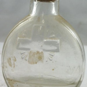 Puede incluir: Una botella de vidrio transparente con tap&oacute;n de corcho. La botella tiene forma redondeada y un dise&ntilde;o de cruz grabado en el vidrio. La botella es probablemente vintage y pudo haber sido utilizada para medicina o perfume.