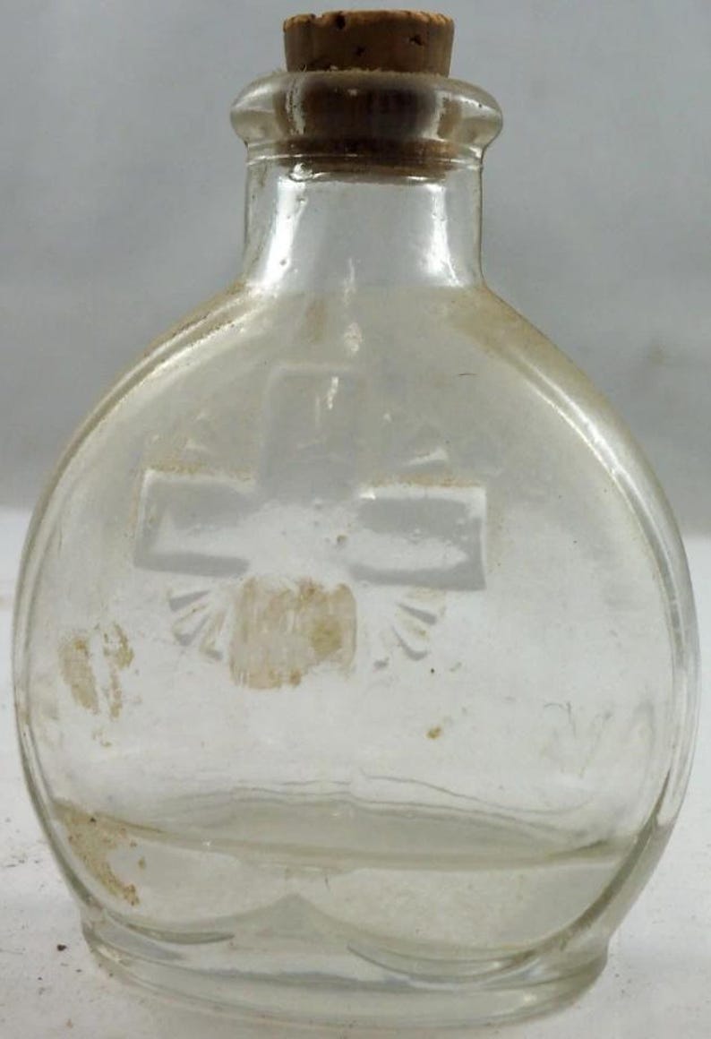 Puede incluir: Una botella de vidrio transparente con tap&oacute;n de corcho. La botella tiene forma redondeada y un dise&ntilde;o de cruz grabado en el vidrio. La botella es probablemente vintage y pudo haber sido utilizada para medicina o perfume.