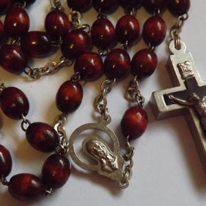 Puede incluir: Un rosario con cuentas de madera de color rojo oscuro y eslabones de cadena de metal plateado. El crucifijo presenta una representaci&oacute;n detallada de Jesucristo. El rosario incluye una pieza central con una figura religiosa.
