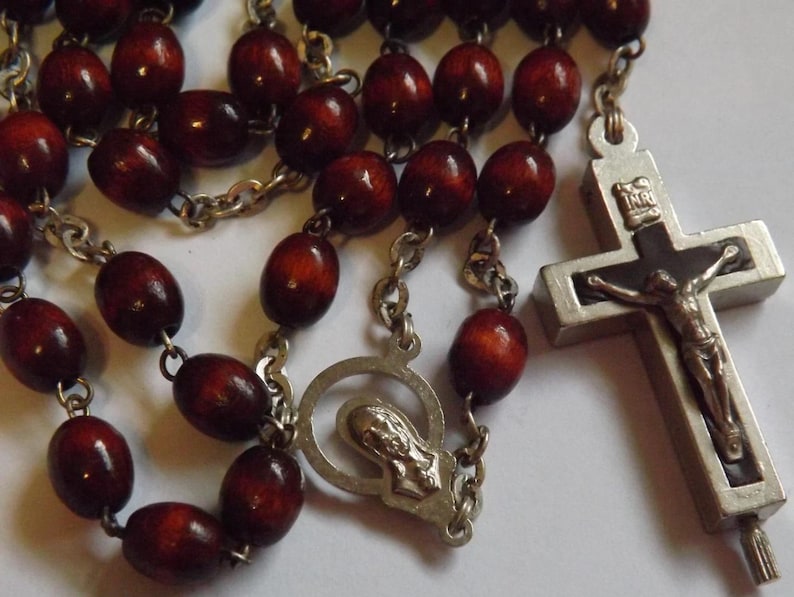 Puede incluir: Un rosario con cuentas de madera de color rojo oscuro y eslabones de cadena de metal plateado. El crucifijo presenta una representaci&oacute;n detallada de Jesucristo. El rosario incluye una pieza central con una figura religiosa.