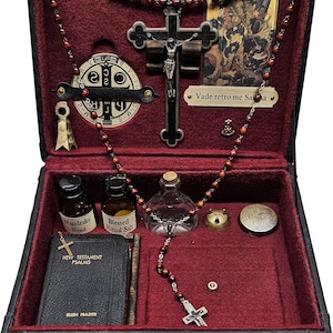 Puede incluir: Una caja negra con interior de terciopelo rojo, que contiene objetos religiosos. Incluye un crucifijo, cuentas de rosario, peque&ntilde;as botellas etiquetadas como "Mandrake Root" y "Blessed Ritual Salt", y un libro titulado "New Testament Psalms" de Ellen Frazier.