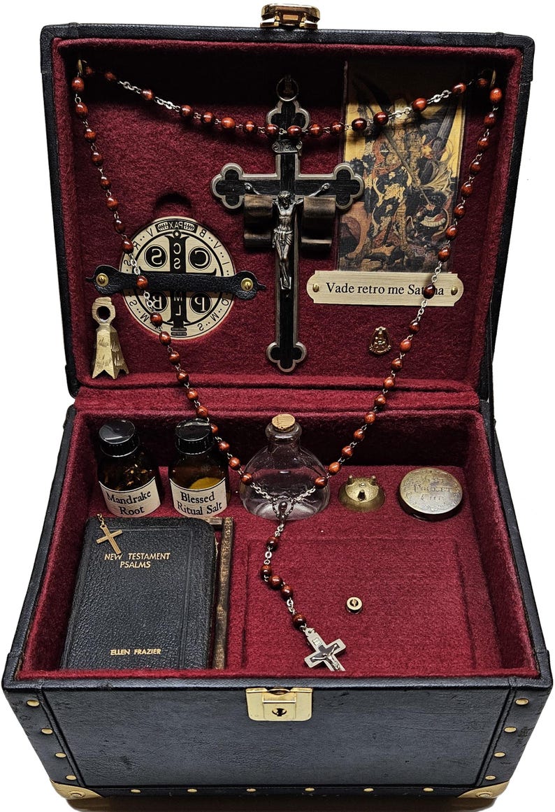 Puede incluir: Una caja negra con interior de terciopelo rojo, que contiene objetos religiosos. Incluye un crucifijo, cuentas de rosario, peque&ntilde;as botellas etiquetadas como "Mandrake Root" y "Blessed Ritual Salt", y un libro titulado "New Testament Psalms" de Ellen Frazier.