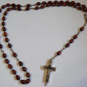 Puede incluir: Un rosario con cuentas ovaladas de color marr&oacute;n oscuro y una cadena de metal plateado. El rosario presenta un crucifijo plateado con una representaci&oacute;n de Jesucristo. Las cuentas est&aacute;n ensartadas, formando un bucle.