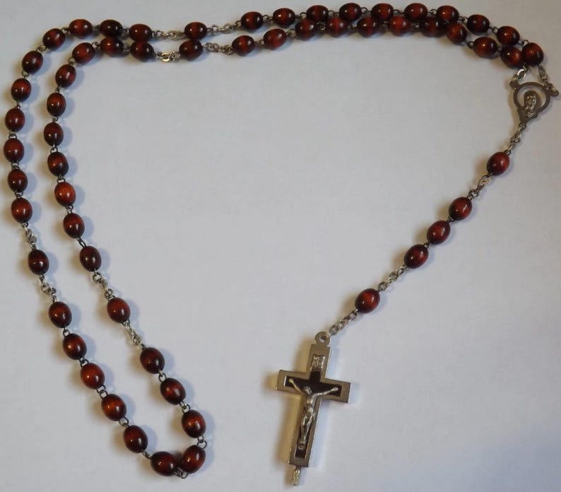 Puede incluir: Un rosario con cuentas ovaladas de color marr&oacute;n oscuro y una cadena de metal plateado. El rosario presenta un crucifijo plateado con una representaci&oacute;n de Jesucristo. Las cuentas est&aacute;n ensartadas, formando un bucle.