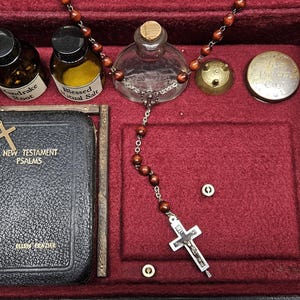 Puede incluir: Una caja forrada de terciopelo rojo oscuro que contiene objetos religiosos. Incluye un peque&ntilde;o libro con una cruz dorada, botellas etiquetadas como "Mandrake Root" y "Blessed Ritual Salt", un rosario y un peque&ntilde;o recipiente de metal. La caja es negra con interior rojo.