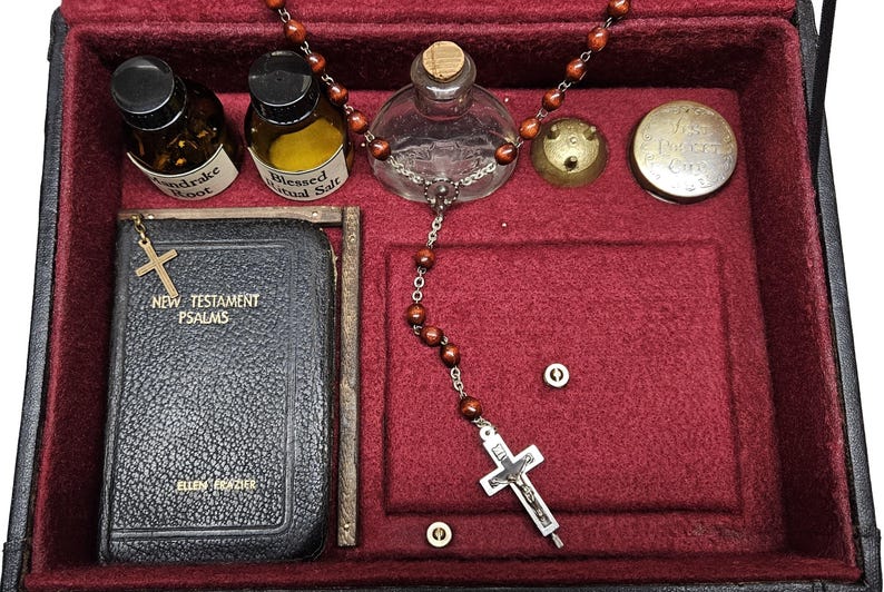 Puede incluir: Una caja forrada de terciopelo rojo oscuro que contiene objetos religiosos. Incluye un peque&ntilde;o libro con una cruz dorada, botellas etiquetadas como "Mandrake Root" y "Blessed Ritual Salt", un rosario y un peque&ntilde;o recipiente de metal. La caja es negra con interior rojo.