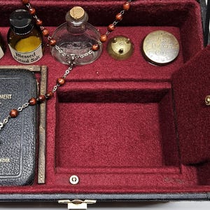 Puede incluir: Una caja negra con interior rojo, que contiene un peque&ntilde;o libro con una cruz, botellas y un rosario. El libro dice "New Testament Psalms". La caja tiene un compartimento con bisagras y un cierre dorado.