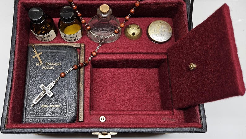 Puede incluir: Una caja negra con interior rojo, que contiene un peque&ntilde;o libro con una cruz, botellas y un rosario. El libro dice "New Testament Psalms". La caja tiene un compartimento con bisagras y un cierre dorado.