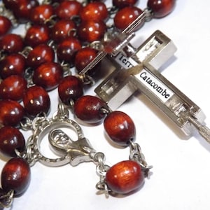 Puede incluir: Un rosario con cuentas de madera marr&oacute;n oscuro y una cruz de metal plateado. La cruz tiene la inscripci&oacute;n "Terra Catacombe". Las cuentas ovaladas est&aacute;n unidas por una cadena plateada.