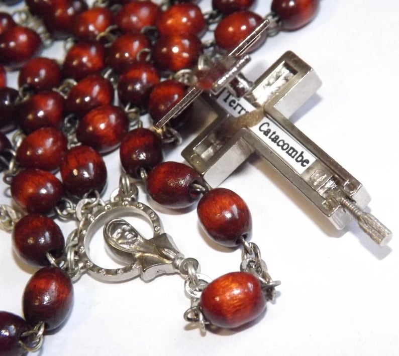 Puede incluir: Un rosario con cuentas de madera marr&oacute;n oscuro y una cruz de metal plateado. La cruz tiene la inscripci&oacute;n "Terra Catacombe". Las cuentas ovaladas est&aacute;n unidas por una cadena plateada.