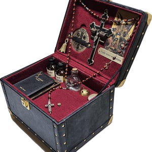 Puede incluir: Un cofre de cuero sint&eacute;tico negro con protectores de esquina dorados y un interior de terciopelo rojo. Contiene objetos religiosos, como un crucifijo, cuentas de rosario, peque&ntilde;as botellas, un libro y una medalla. El cofre est&aacute; abierto, mostrando su contenido.