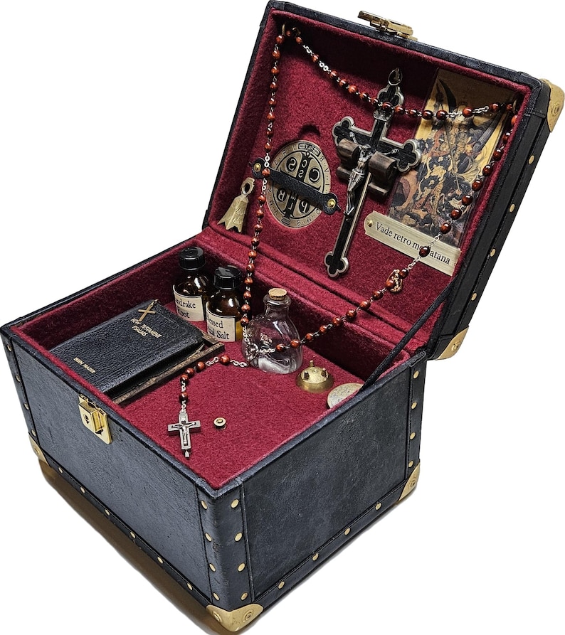 Puede incluir: Un cofre de cuero sint&eacute;tico negro con protectores de esquina dorados y un interior de terciopelo rojo. Contiene objetos religiosos, como un crucifijo, cuentas de rosario, peque&ntilde;as botellas, un libro y una medalla. El cofre est&aacute; abierto, mostrando su contenido.