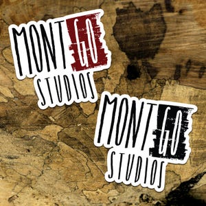 May include: Two white stickers with black text that reads "MONTGO STUDIOS". One sticker has a red "GO" and the other has a black "GO". The stickers are on a brown wooden surface.