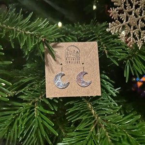 May include: Silver crescent moon earrings with a hammered texture, hanging from a card with the text "Wildwood Jewellery".