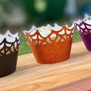 May include: Three cupcakes with white frosting and spiderweb cupcake wrappers. The wrappers are black, orange, and purple.