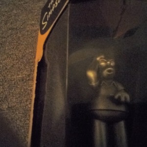 May include: A black and gray figurine of a cartoon character with a yellow light bulb in its hand. The figurine is in a box with the word "The Simpsons" printed on it.