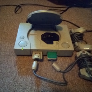 May include: A white PlayStation console with the top open, revealing the disc drive. Two gray controllers are connected to the console with cords.