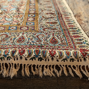Può includere: Primo piano di un tessuto decorativo con un motivo paisley dettagliato in rosso, blu e oro su sfondo bianco sporco. Il bordo presenta una frangia di nappe color crema. Il tessuto è probabilmente un tappeto o un arazzo.
