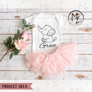 May include: A white onesie with a black outline of an elephant and the name "Grace" written underneath. The onesie is laying on a wooden surface with a pink tulle skirt and pink roses.