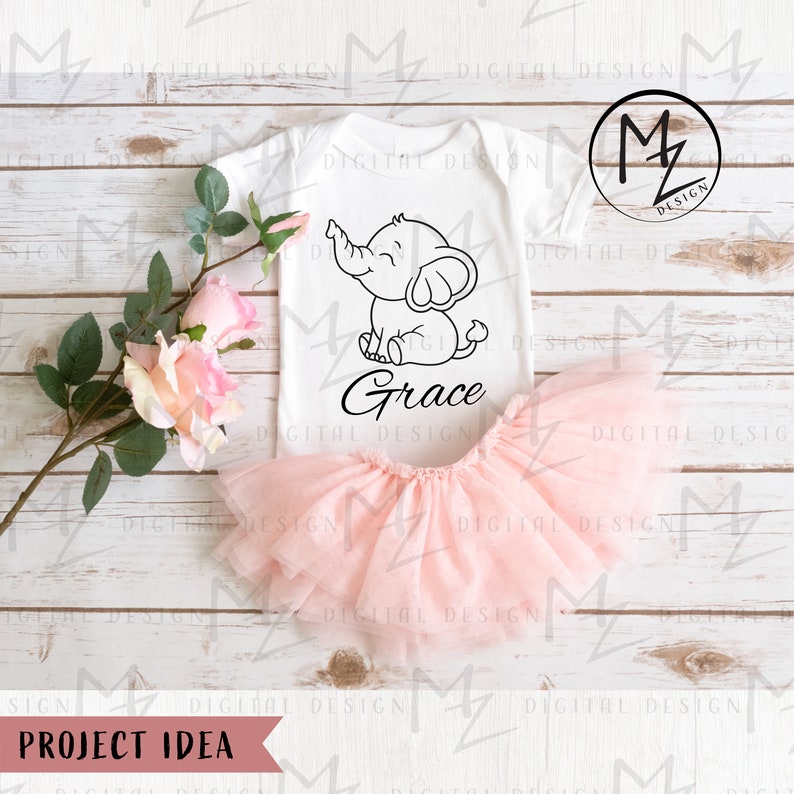 May include: A white onesie with a black outline of an elephant and the name "Grace" written underneath. The onesie is laying on a wooden surface with a pink tulle skirt and pink roses.