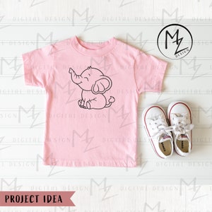May include: A pink t-shirt with a black line drawing of an elephant on the front. The elephant is smiling and has its trunk raised. The t-shirt is laying on a white wooden surface with a pair of white sneakers.