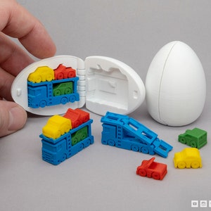 May include: A white egg-shaped container opens to reveal a blue toy truck carrying smaller toy cars in red, yellow, and green. The truck has a ramp that can be lowered. There are also several individual toy cars in red, yellow, and green.