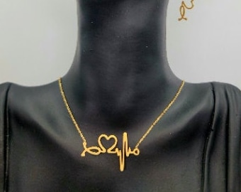 May include: Gold necklace with a stethoscope and heartbeat design. The necklace is worn by a model with gold earrings featuring a heart and cross design.
