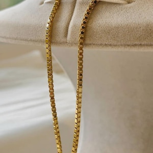 May include: A gold-colored box chain necklace. The chain is displayed against a neutral-toned background. The necklace is made of small, rectangular links that create a smooth, continuous surface. The chain is a classic jewelry piece.