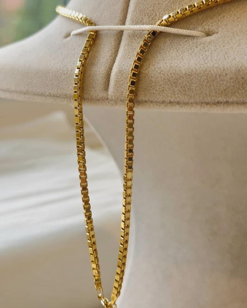 May include: A gold-colored box chain necklace. The chain is displayed against a neutral-toned background. The necklace is made of small, rectangular links that create a smooth, continuous surface. The chain is a classic jewelry piece.